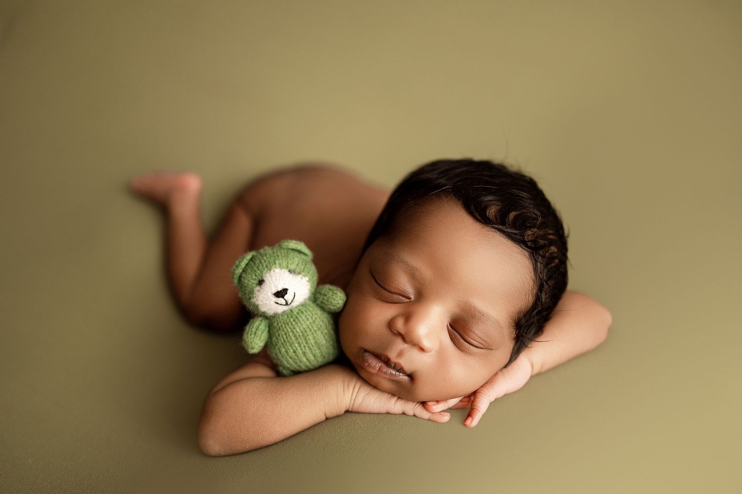 Sleeping newborn baby resting chin on hands beside a small green knitted teddy bear on olive background.