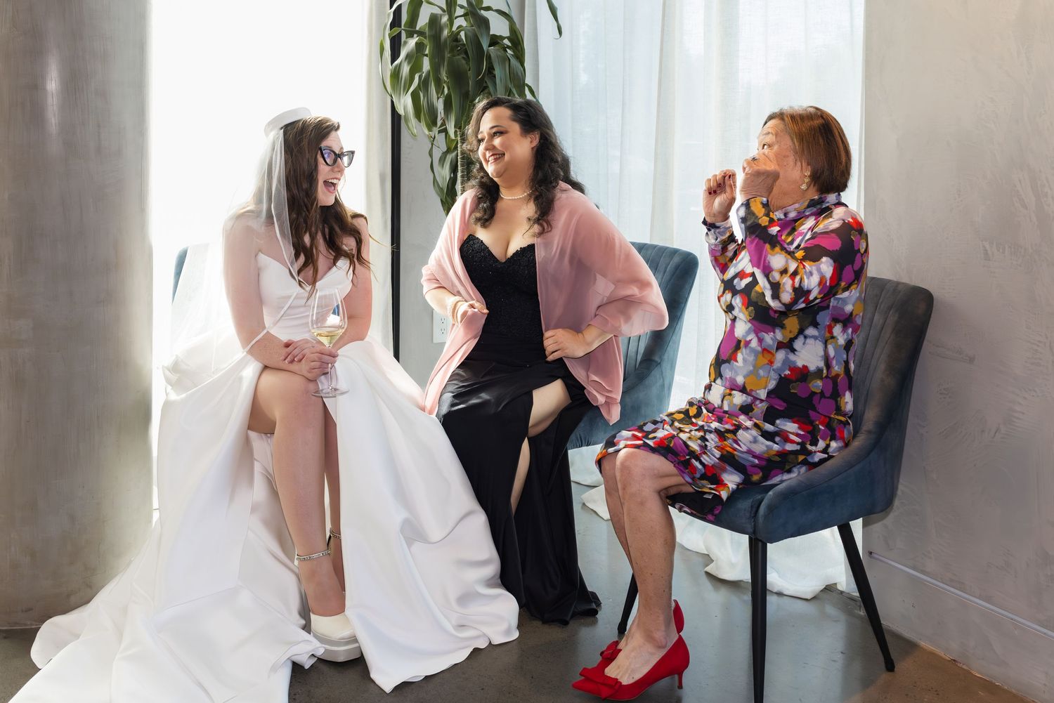 Bride in white gown sits with two women, one in black dress and one in floral outfit, sharing a joyful moment before wedding.