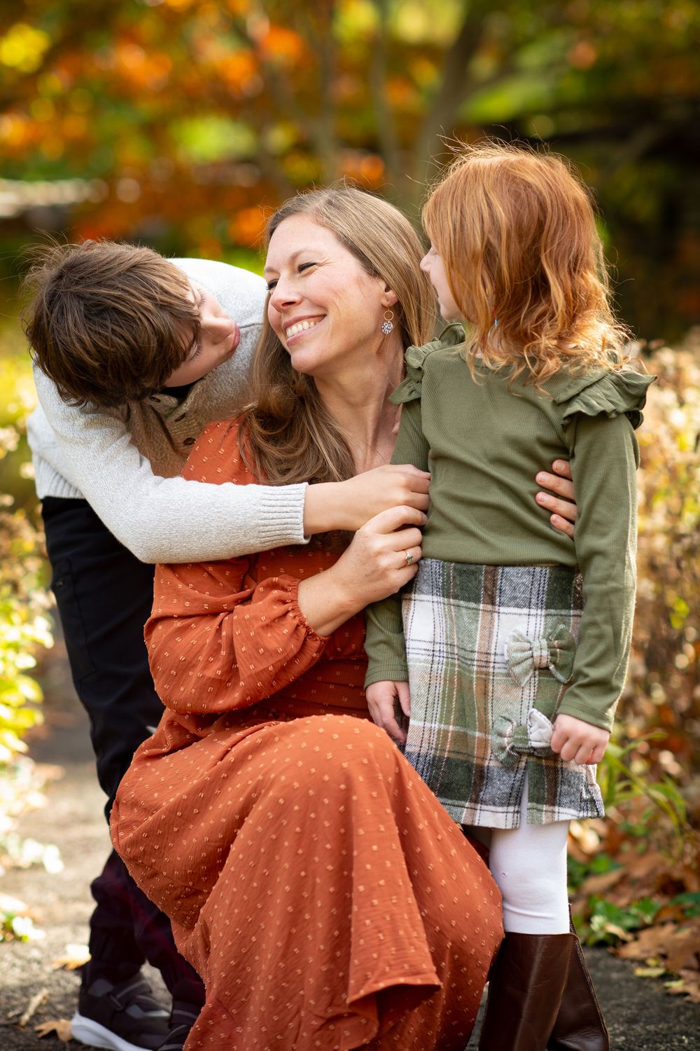 A smiling mother in an orange dress embraces her red-haired daughter and son during a fall family portrait outdoors.