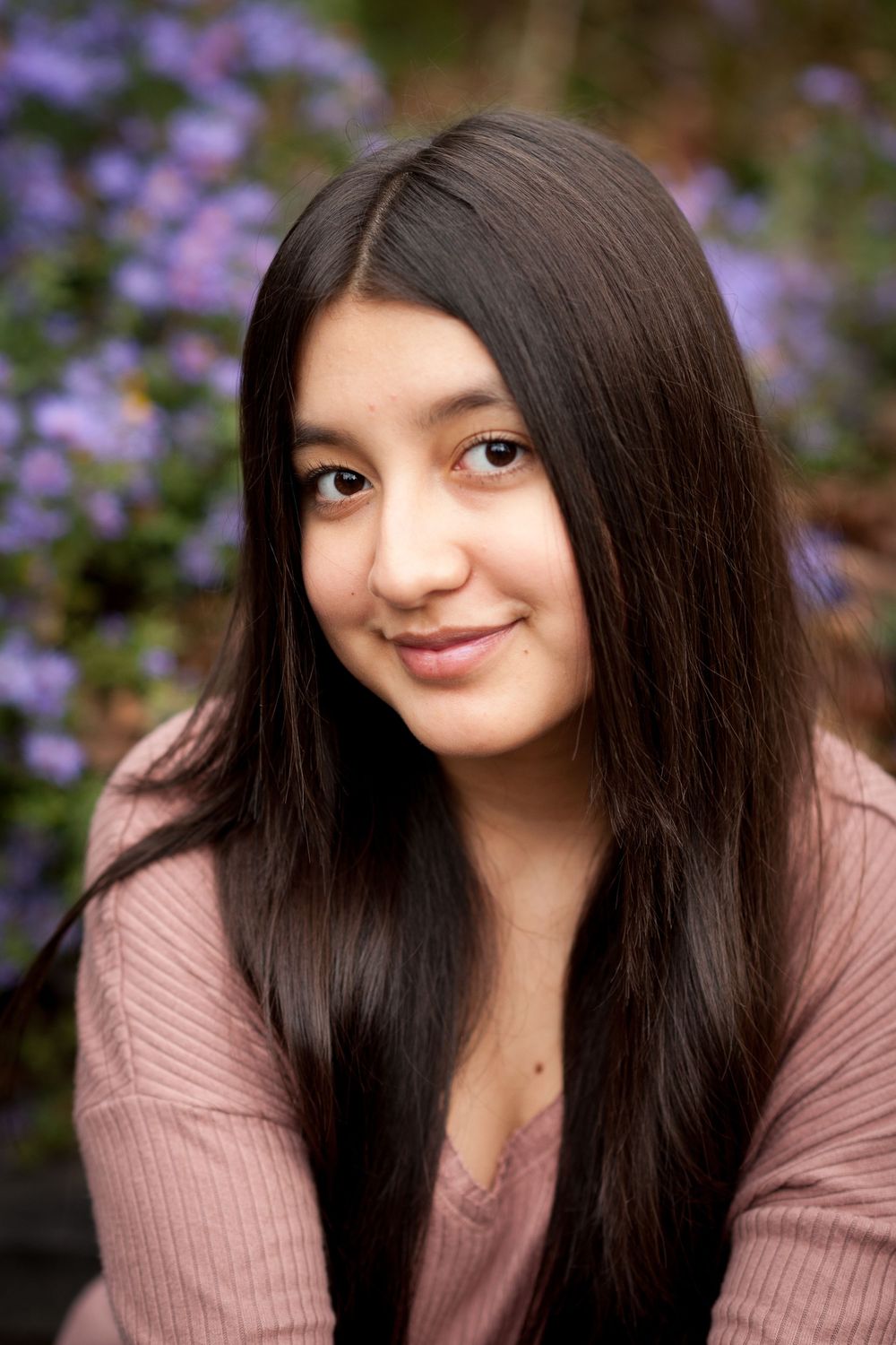 Young woman with long dark hair smiling softly outdoors, wearing a pink top with purple flowers in the background.