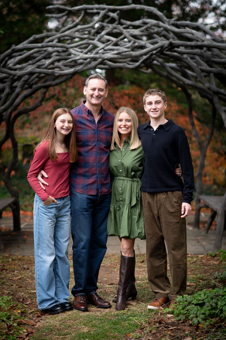 Family of four smiling outdoors under rustic branch archway during fall season in a garden setting.