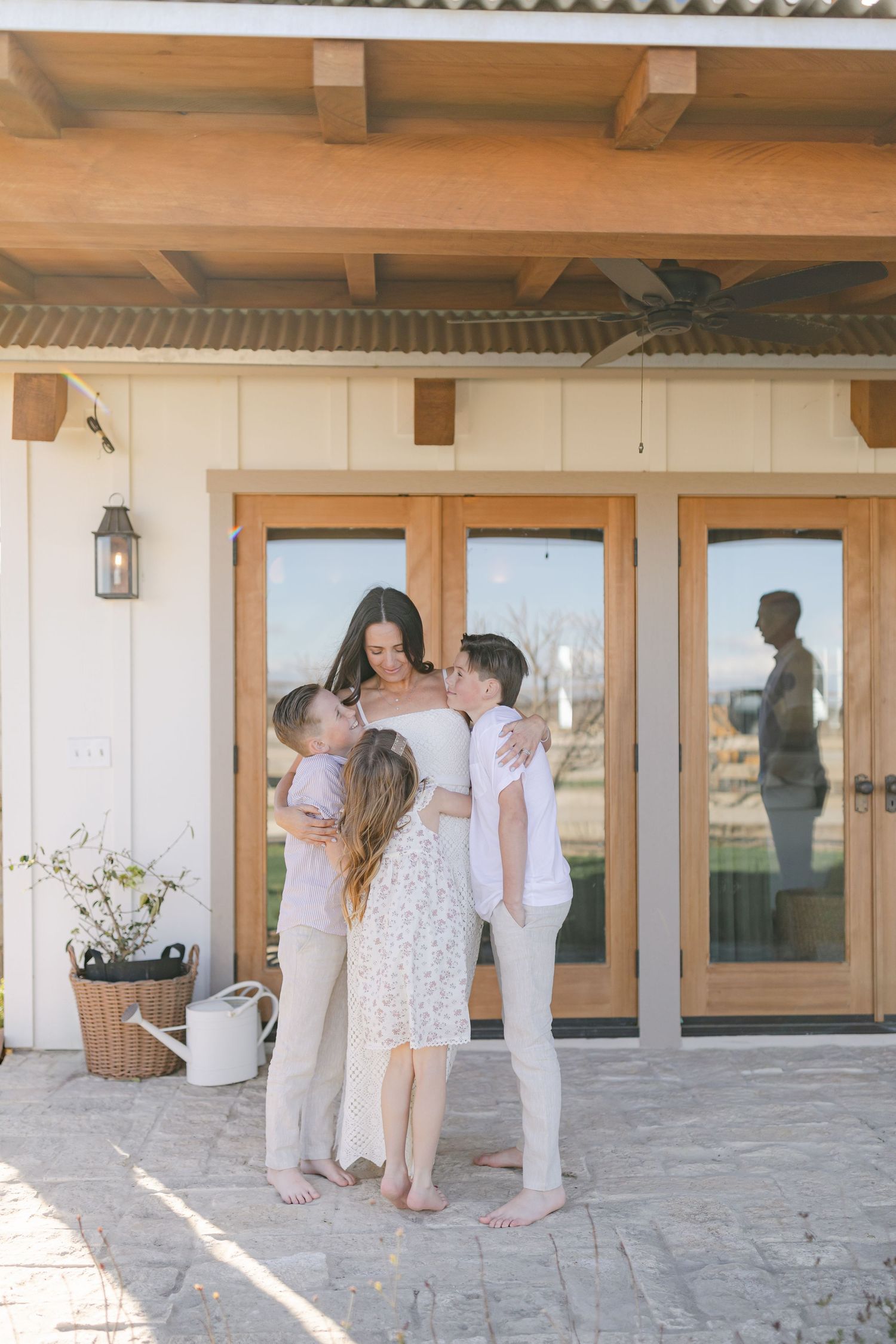 A mother embraces her three children on a sunny patio outside a modern farmhouse with French doors.