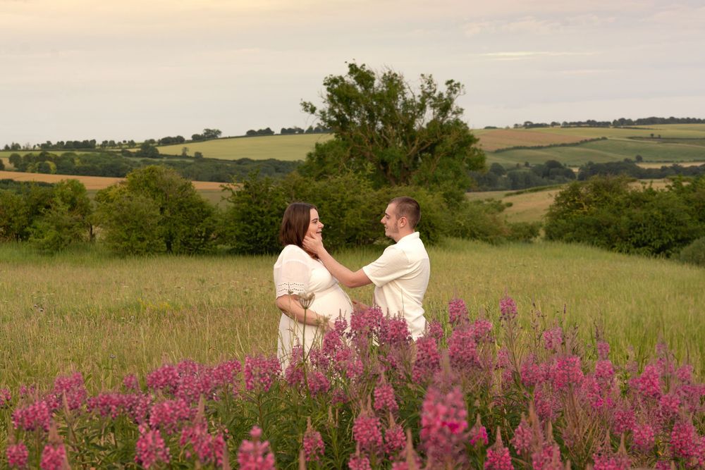 Couple holding hands in a wildflower meadow at sunset, surrounded by pink flowers and rolling green hills.