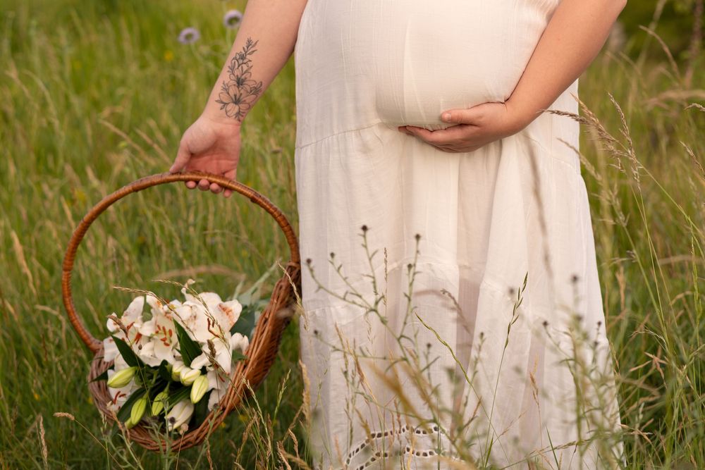 Pregnant woman in white dress holding a flower basket in a green field, showcasing a floral arm tattoo.