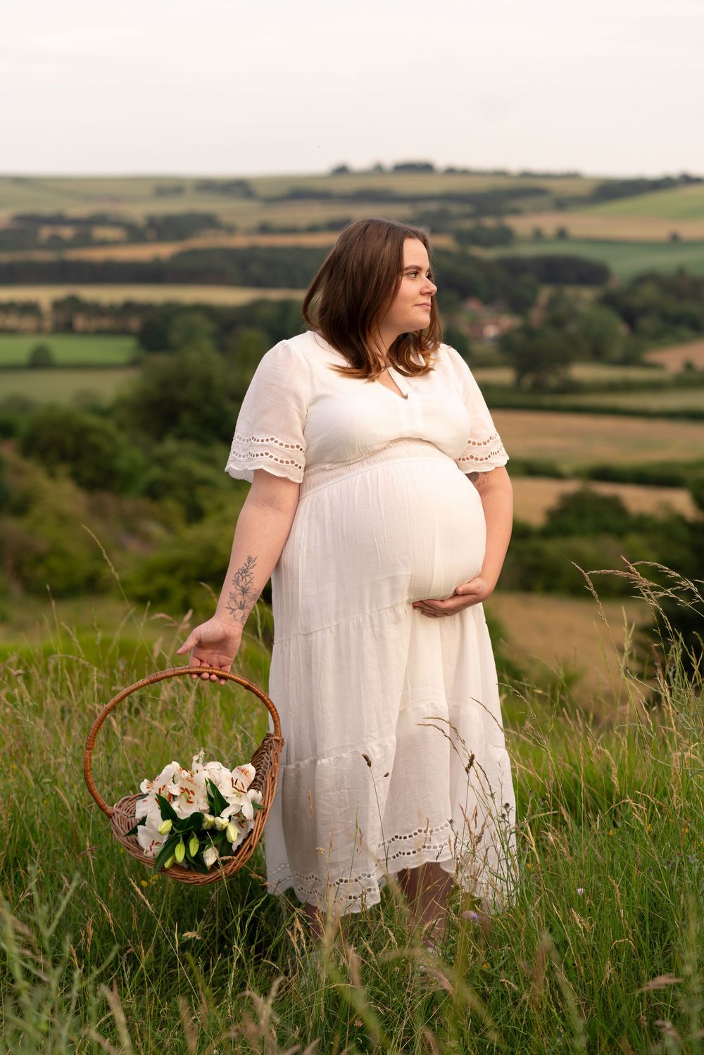 Pregnant woman in white dress holding flower basket in scenic green countryside.
