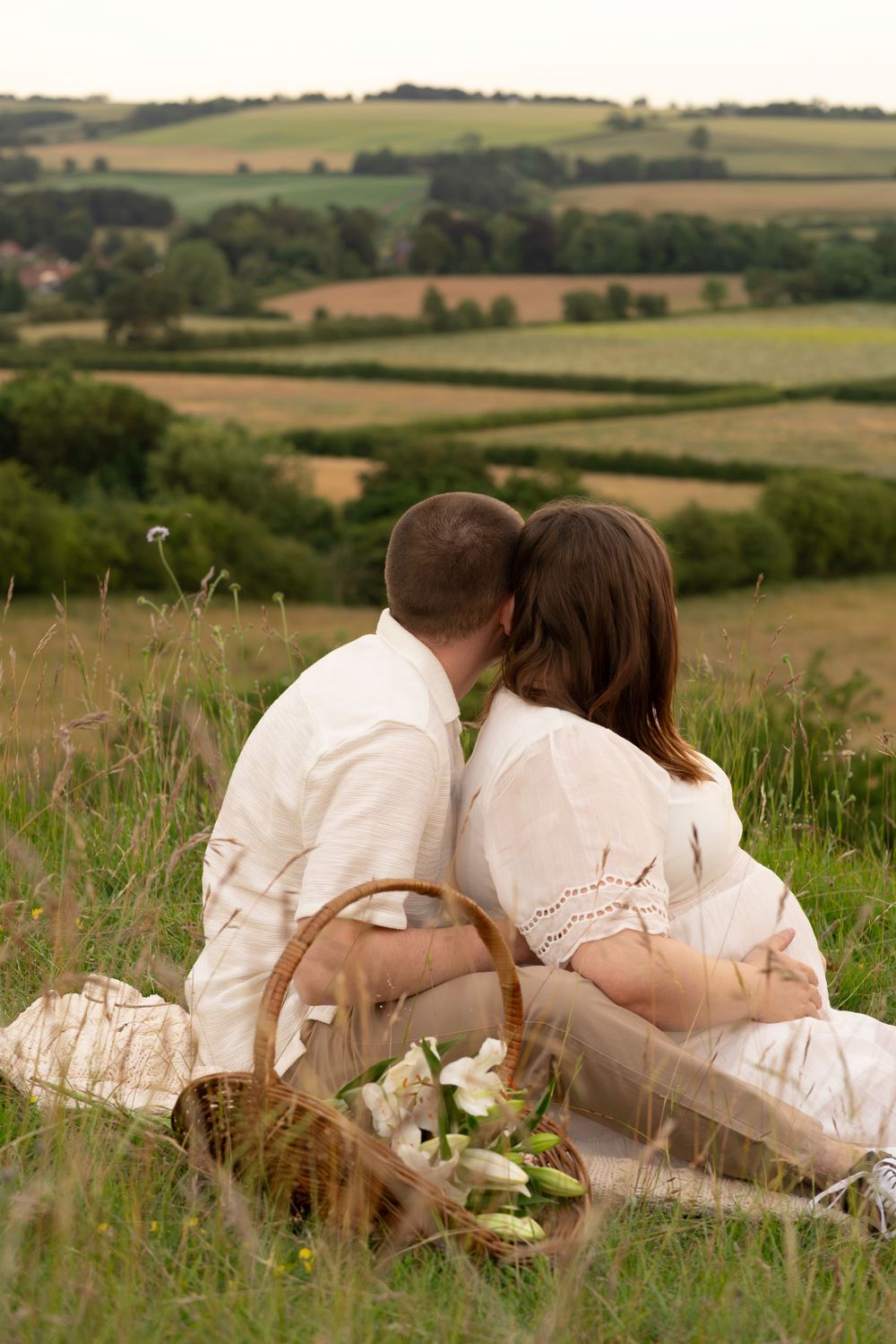 Couple sitting in grass field kissing with flower basket and rolling hills behind.