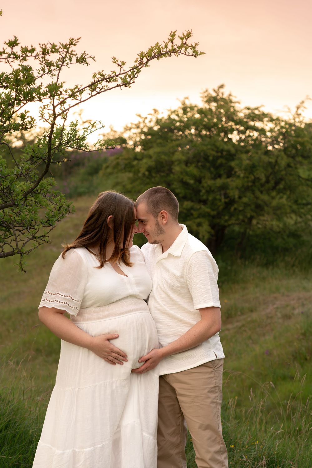 Expecting couple touching baby bump tenderly at sunset in lush green landscape.