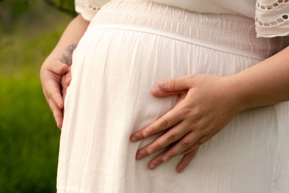 Close-up of a pregnant woman in a white dress gently cradling her baby bump with both hands outdoors.