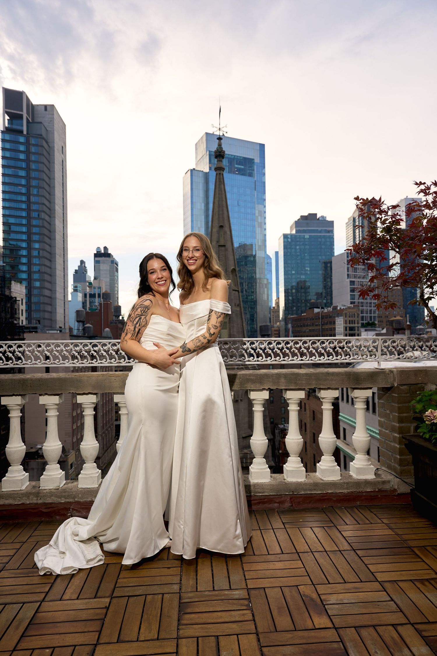 Two brides in white gowns embrace on a rooftop terrace with NYC skyline backdrop on their wedding day.