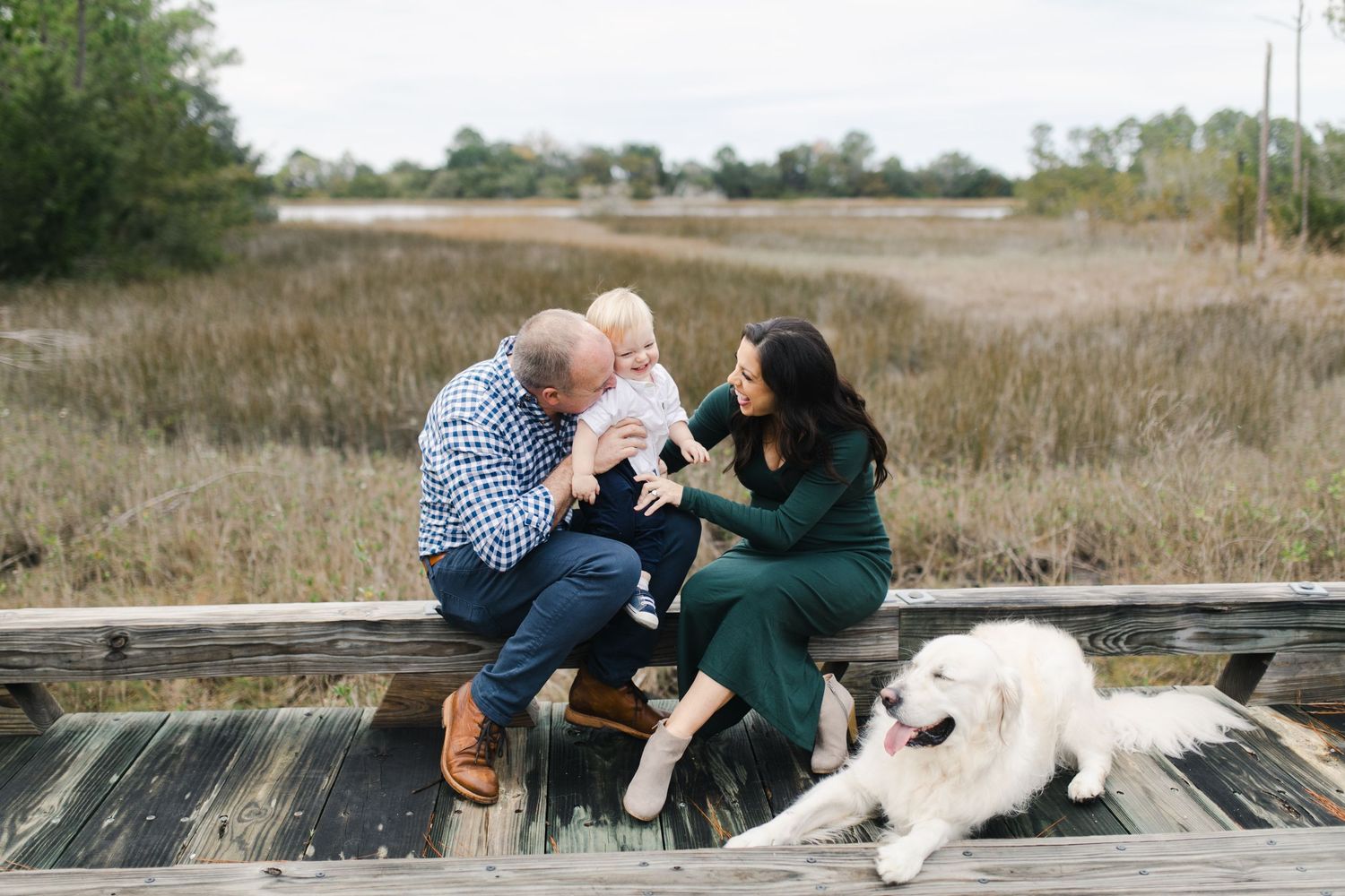 Family with baby and white dog sitting on wooden dock surrounded by marshland during outdoor photo session.