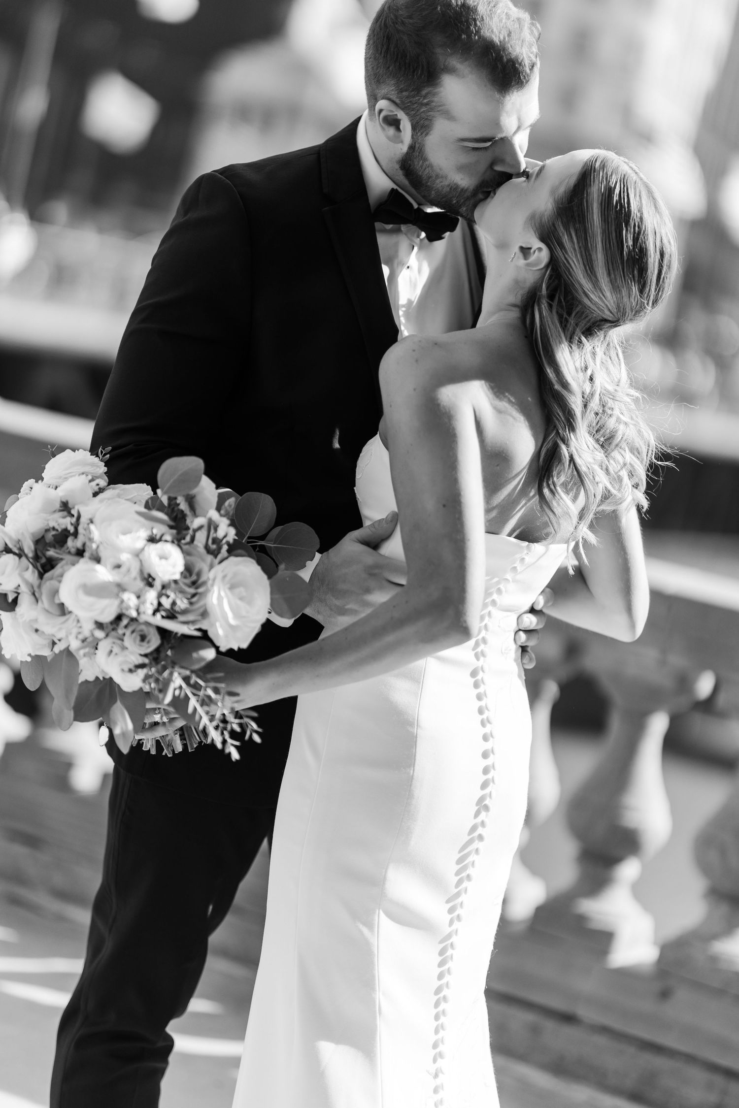 Black and white photo of a groom in a tuxedo kissing his bride in a white gown while holding a floral bouquet.