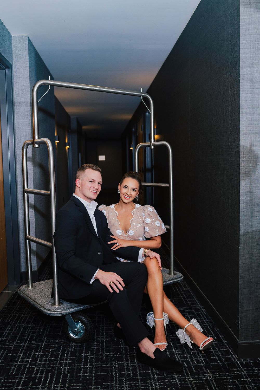 Elegant couple in formal attire sitting on a hotel luggage cart in a stylish dark corridor.