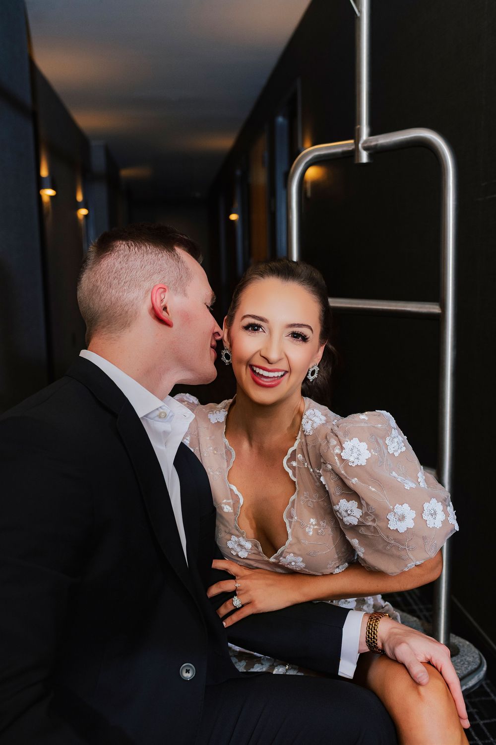 Couple in elegant attire sharing a romantic moment in a dimly lit hallway, woman smiling in floral dress.