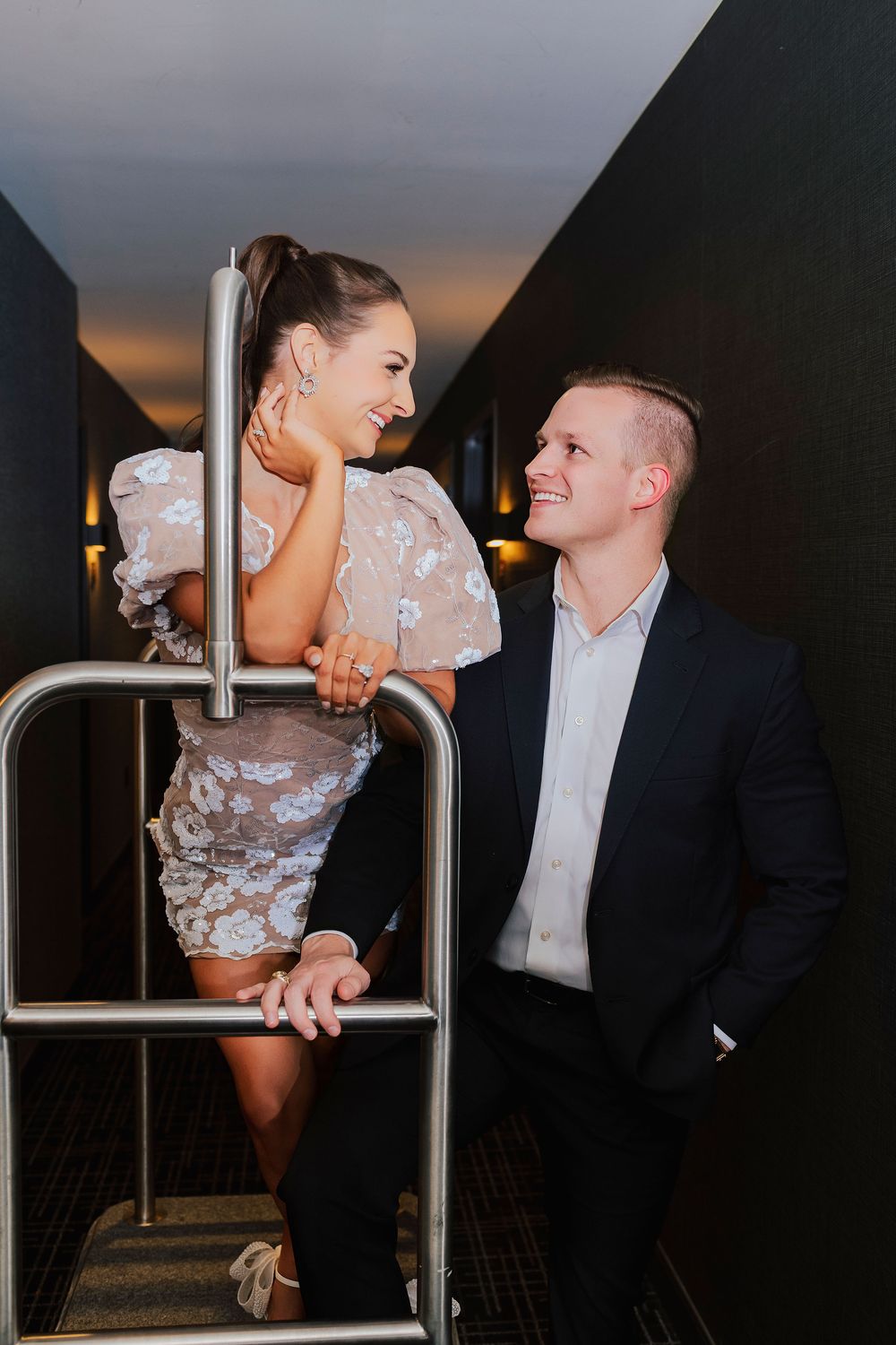 A smiling couple poses on a staircase railing, woman in floral mini dress, man in dark suit, sharing a joyful moment.