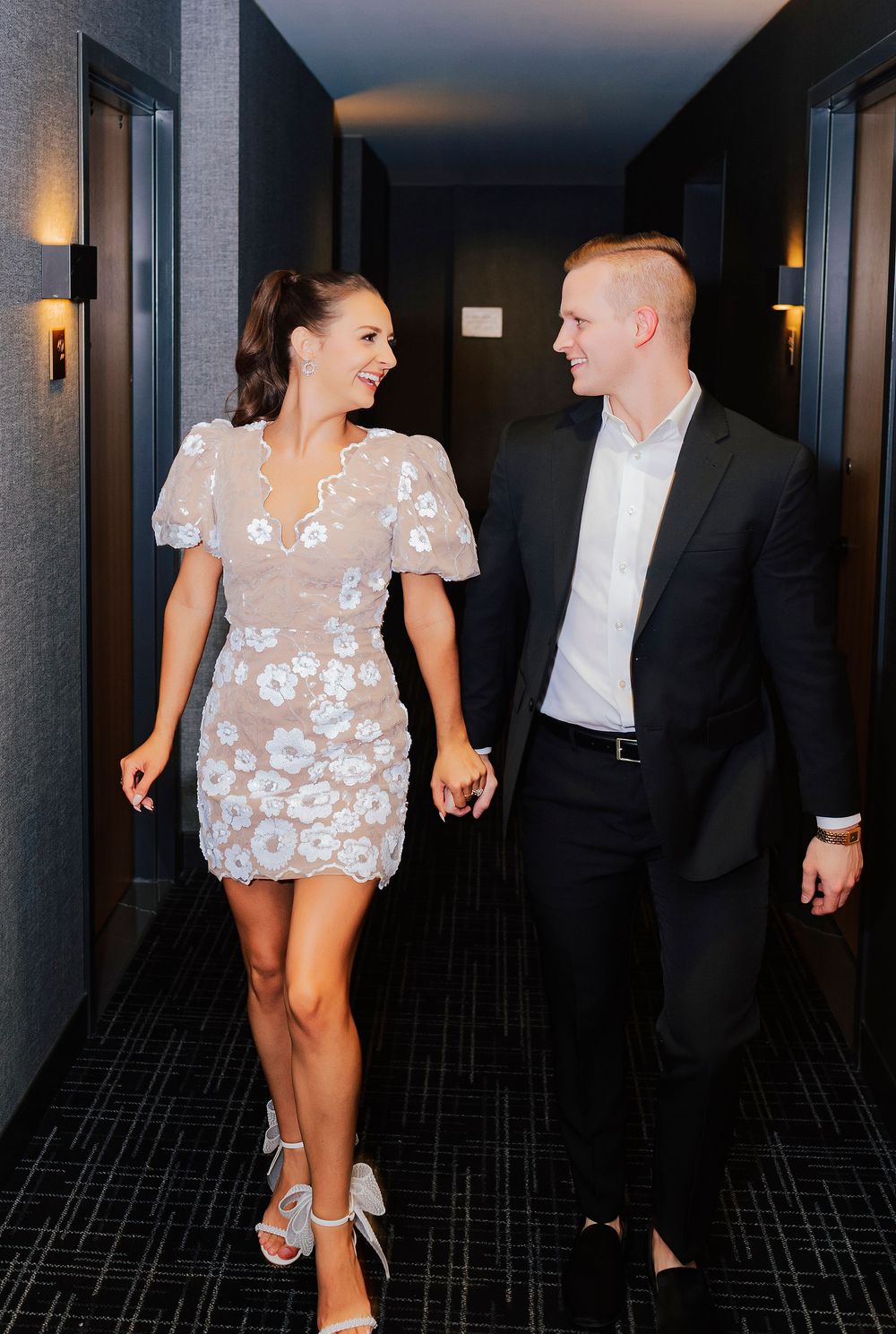 Couple walking hand in hand down a hotel hallway, woman in floral mini dress and man in black suit.