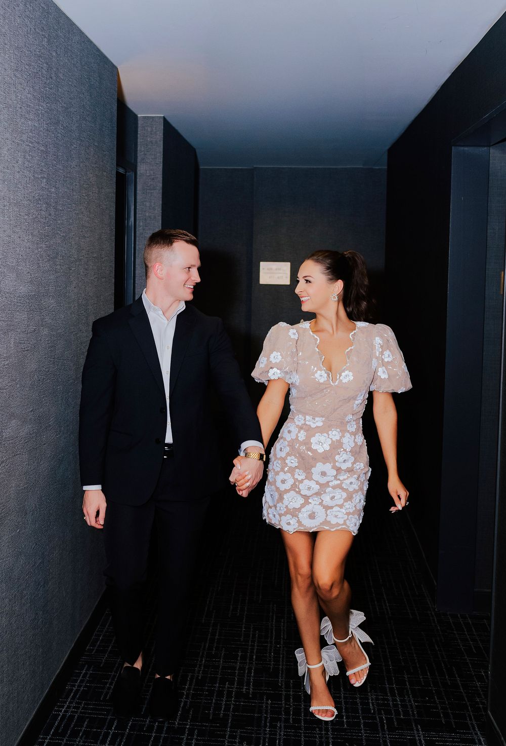 Couple holding hands walking down a hallway, woman in floral mini dress and heels, man in black suit.
