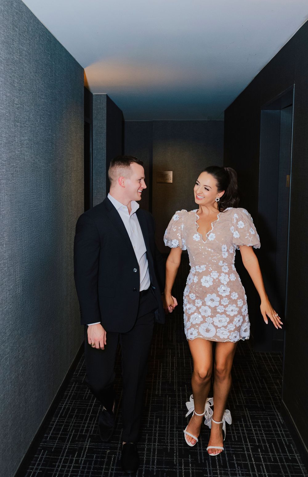 Couple walking hand in hand down a modern hallway, woman in floral mini dress and man in navy suit smiling at each other.