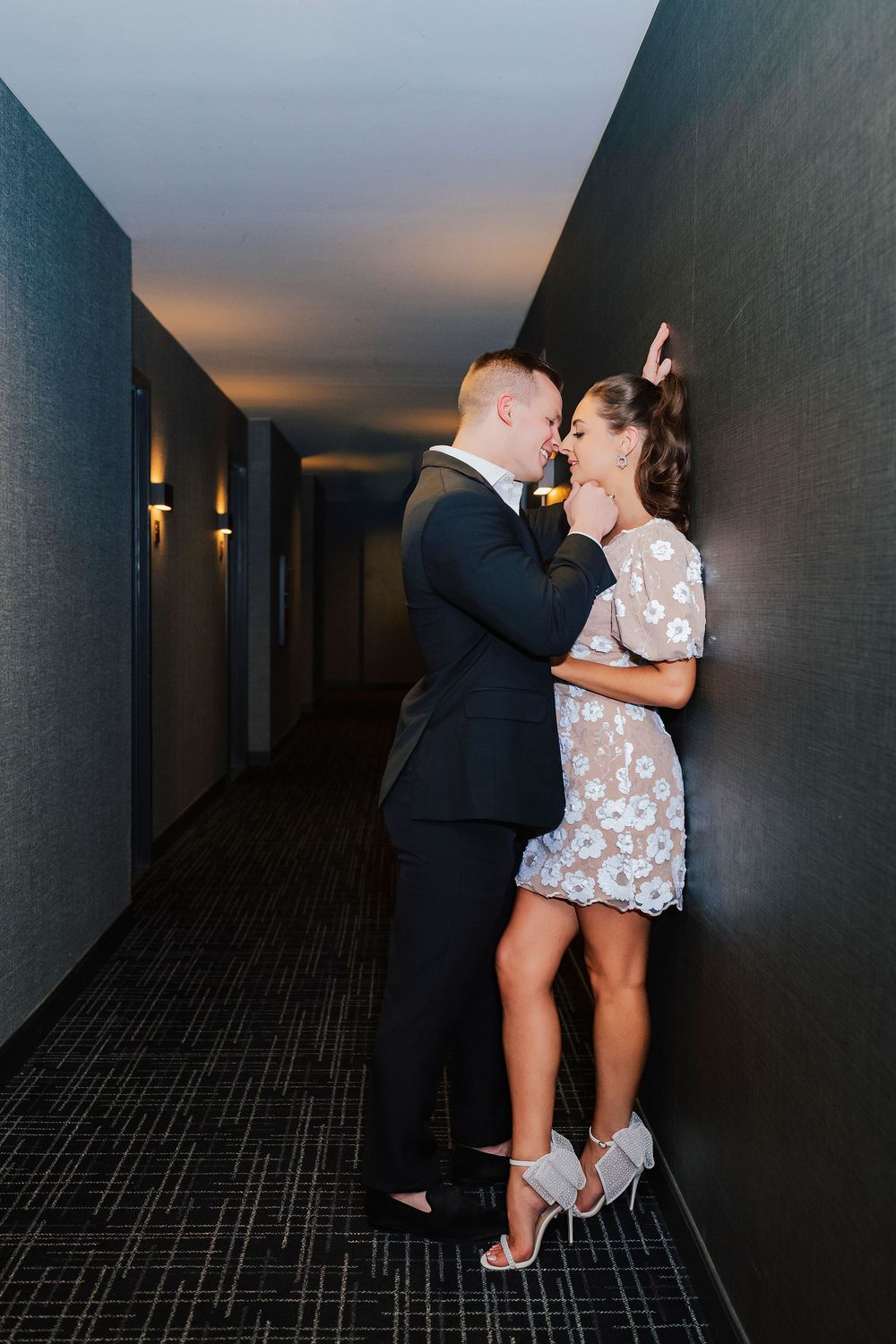 Couple sharing a romantic moment in a dimly lit hotel hallway, man in suit and woman in floral mini dress and heels.