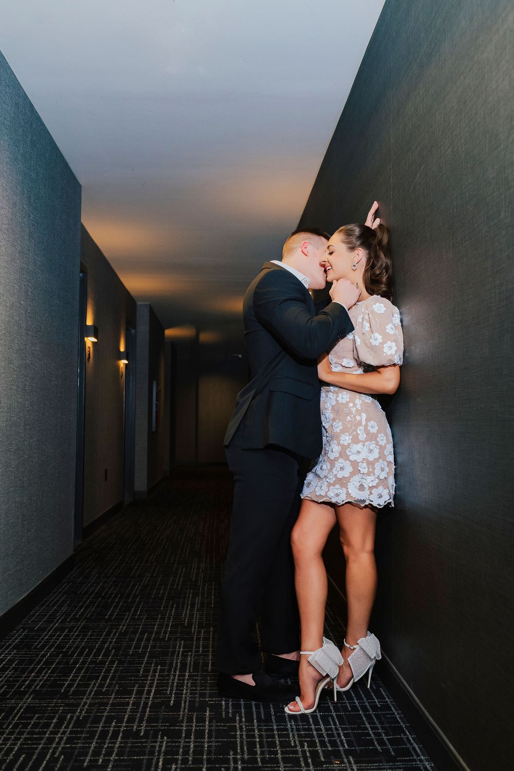 Couple sharing a romantic kiss in a dimly lit hotel corridor, woman in white lace dress and heels, man in dark suit.