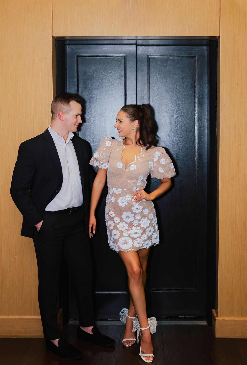 Stylish couple smiling at each other in front of a black door, woman in lace floral mini dress and heels.