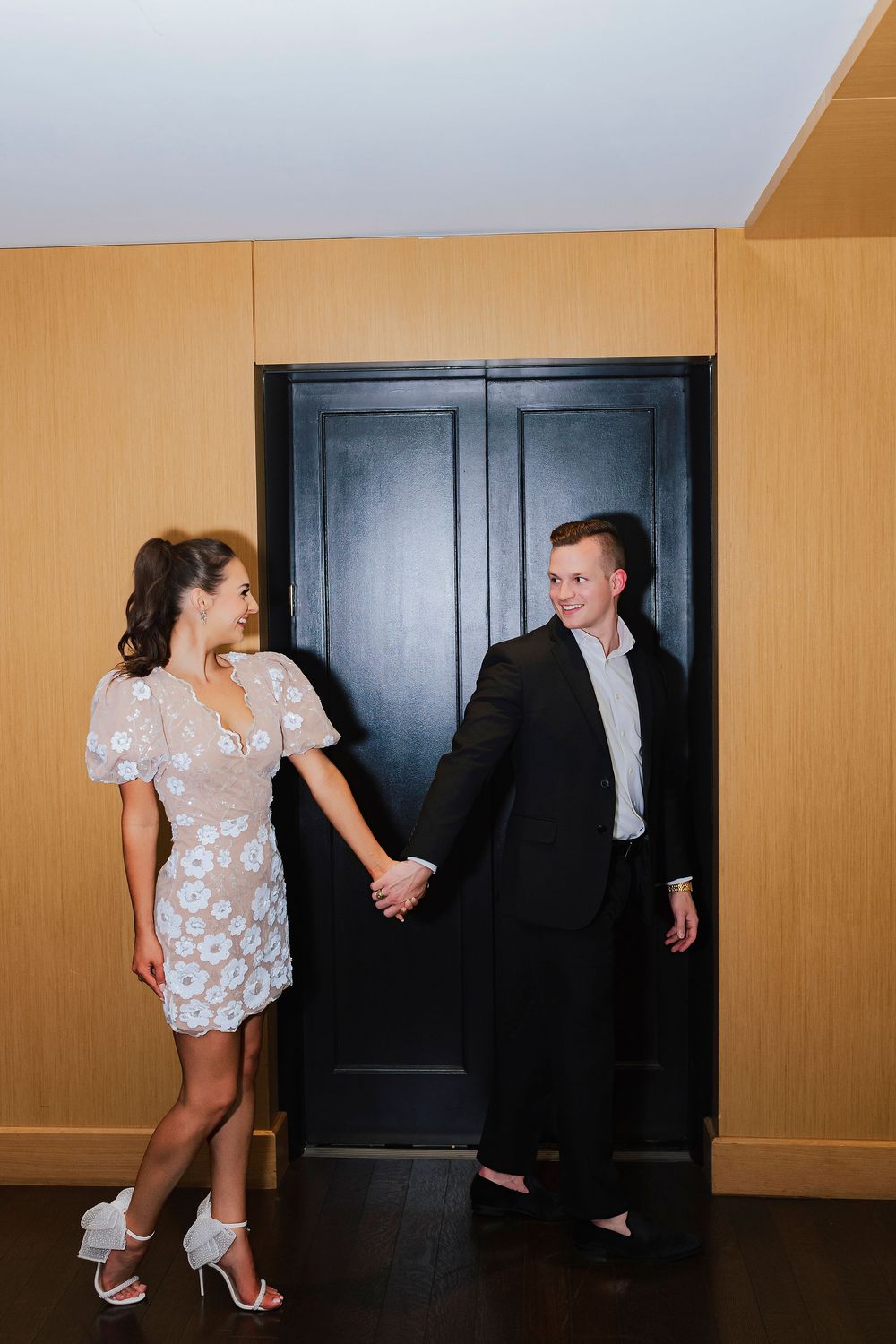 Elegant couple holding hands by a black door, woman in floral mini dress and heels, man in black suit.
