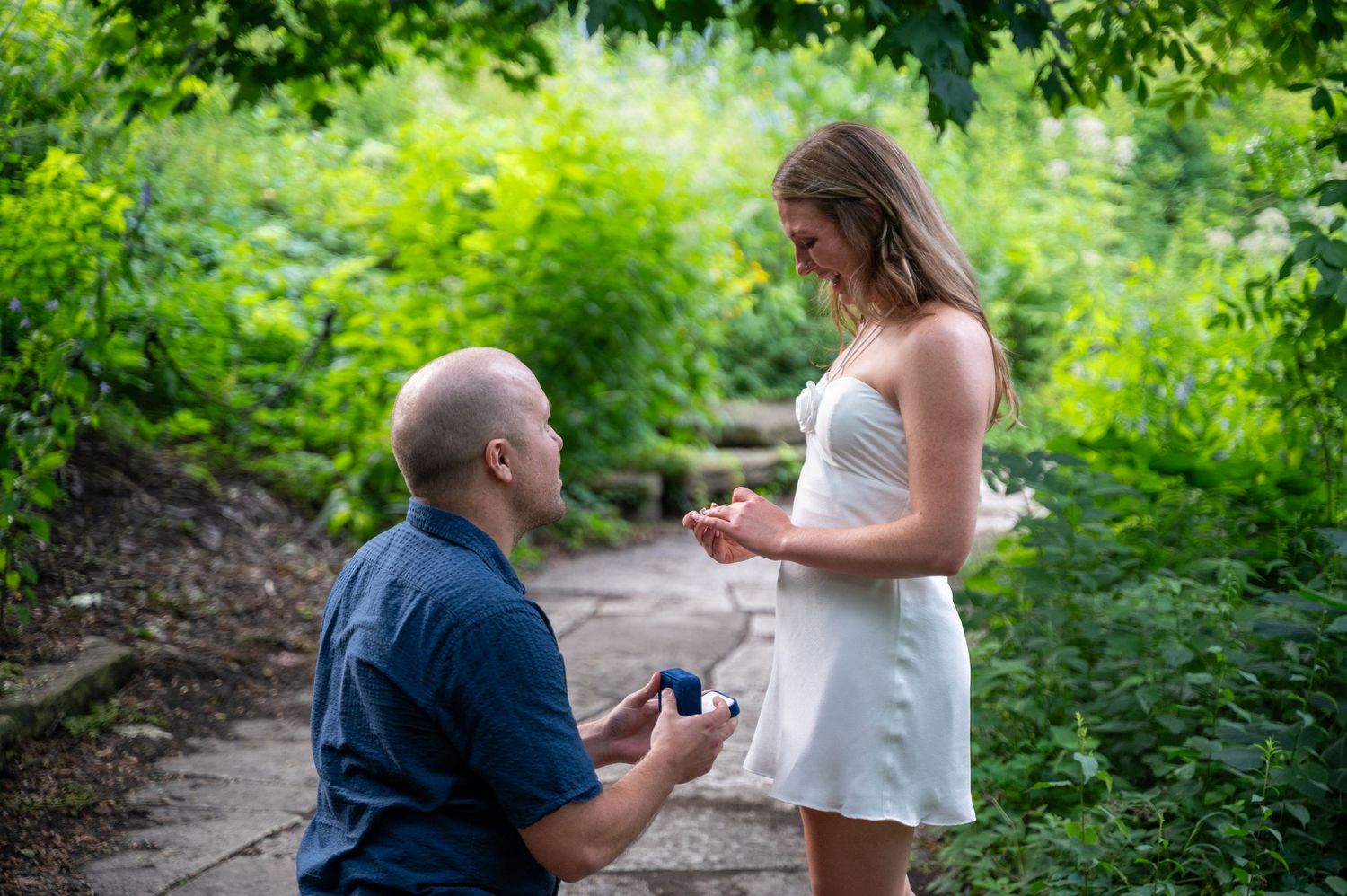Man kneeling on one knee proposing to woman in white dress outdoors in lush green park setting.