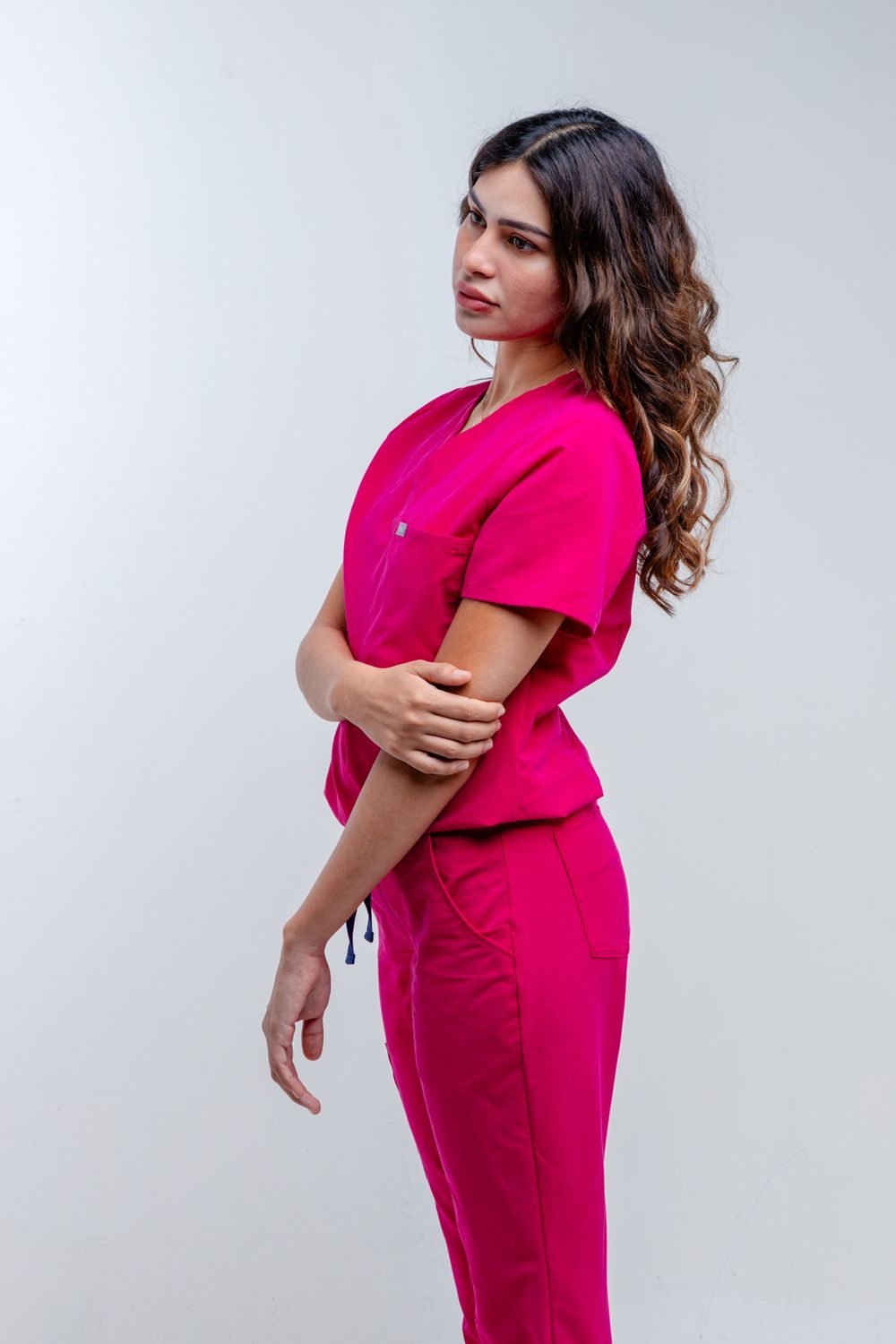 Woman in pink medical scrubs posing against white background, holding her elbow with curly dark hair.