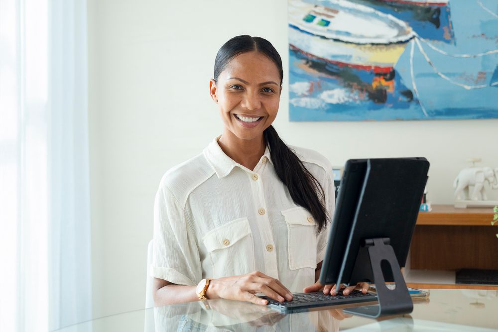 Smiling woman working on tablet at glass desk with colorful boat painting on wall behind her.