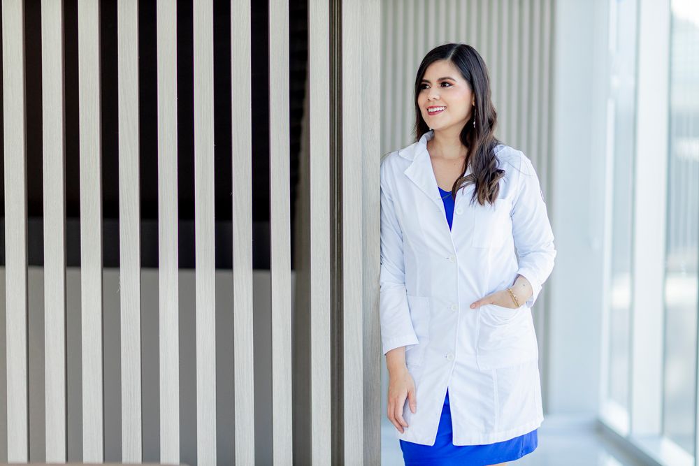 A smiling female doctor in a white lab coat and blue scrubs stands confidently in a modern medical facility.