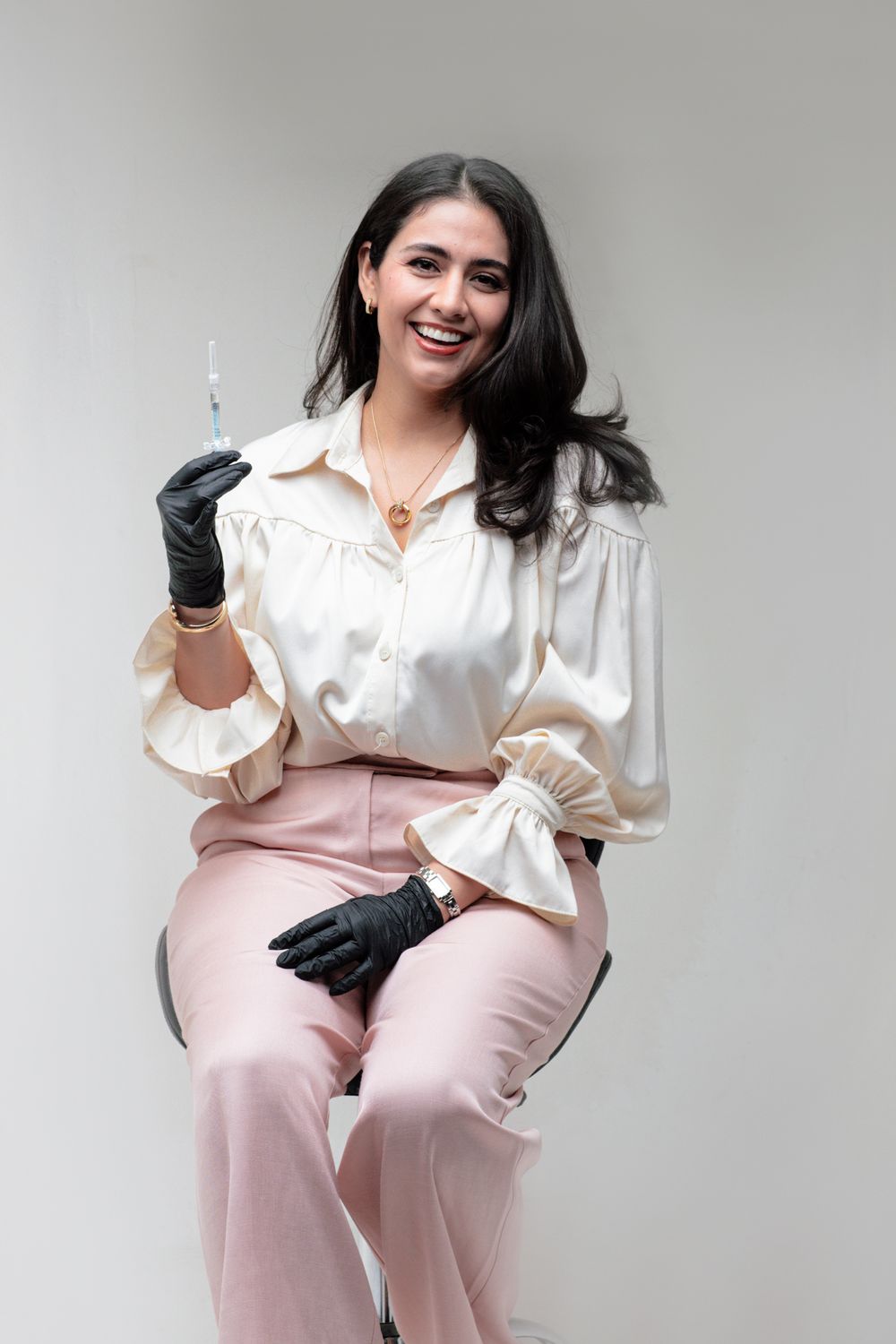 Smiling woman in white blouse and pink pants wearing black gloves holds a syringe while seated against a light background.