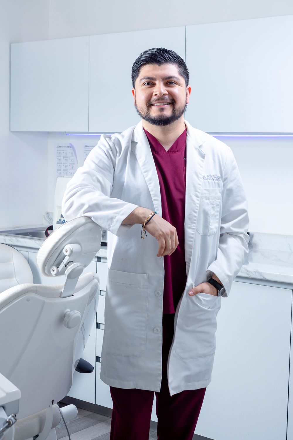 A smiling male dentist in a white lab coat and burgundy scrubs poses confidently in a modern dental clinic.
