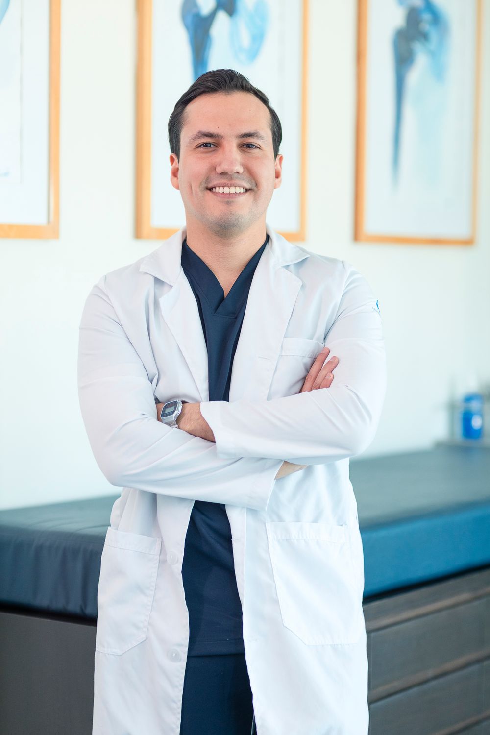Smiling male doctor in white lab coat with arms crossed standing in medical office with anatomical charts.