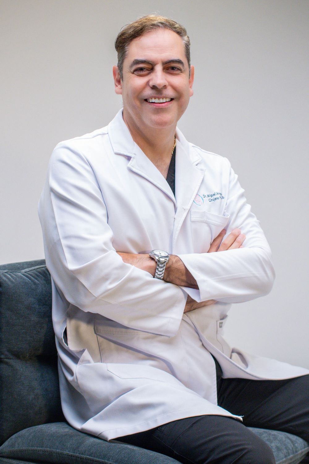 Smiling male doctor in white lab coat sitting with arms crossed in a medical office setting.