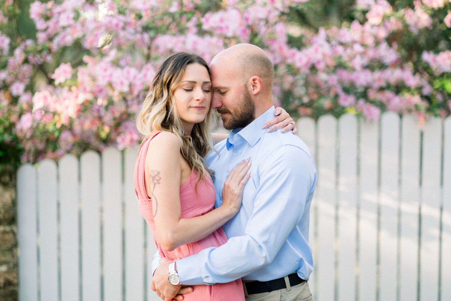 Engagement photography in Savannah, GA of couple embracing in front of white fence with pink blossoming flowers, woman in pink, man in blue.