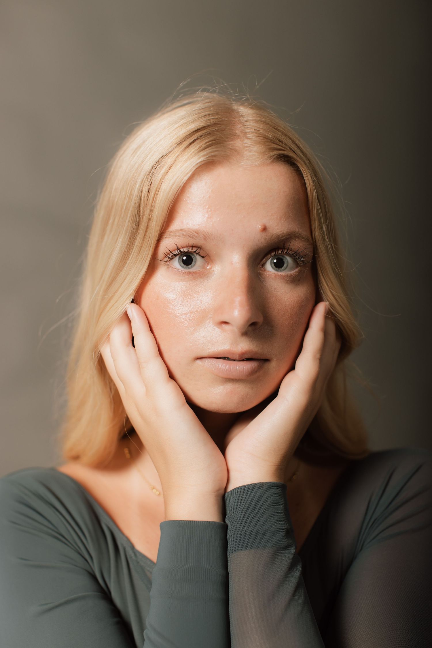 Young blonde woman with blue eyes holds her face in her hands, wearing sage green against a gray background for in-studio dance photography.