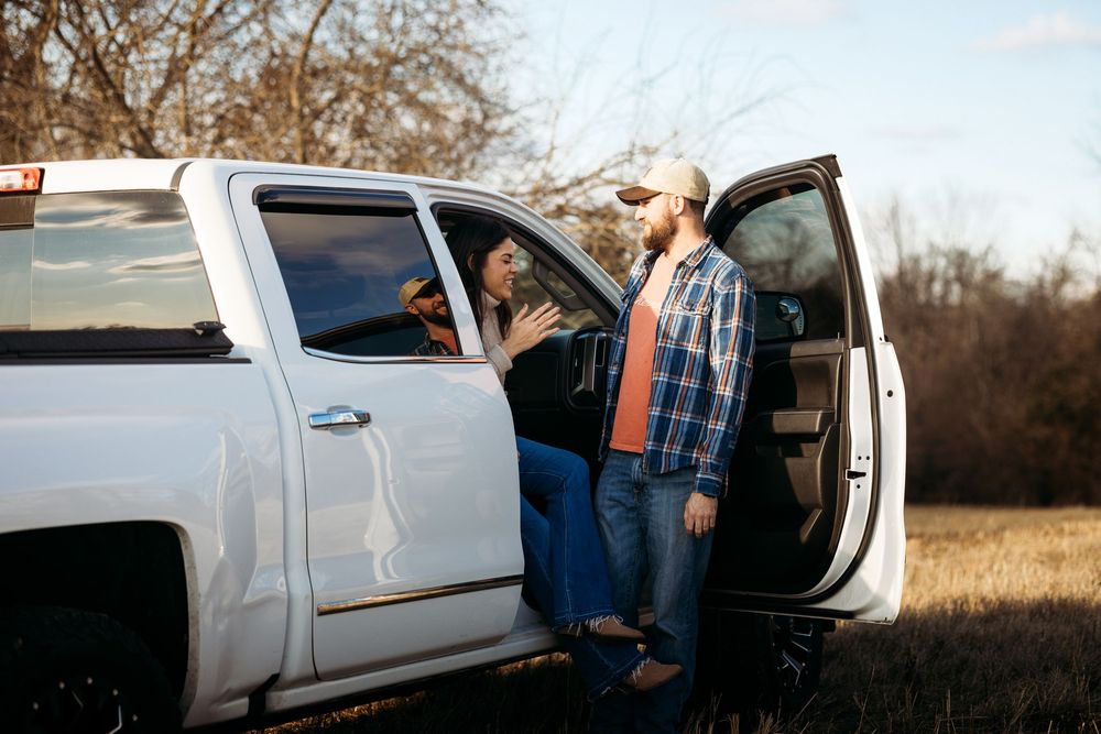 A man and woman chat beside an open white pickup truck door in a rural field at golden hour.