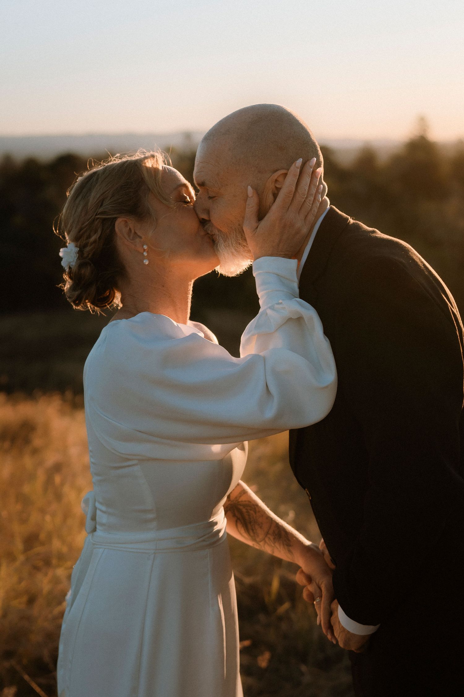 Mature bride and groom sharing a romantic kiss at golden hour sunset, bride in white dress with floral hair accessory.