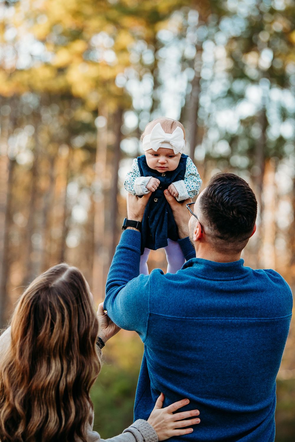 Parents lifting their baby girl up in a sunlit forest during a family photo session.