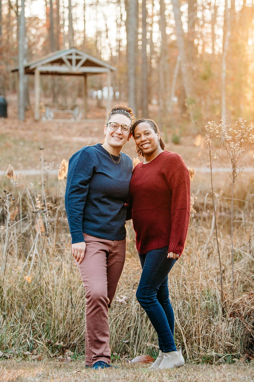 Two women smiling and embracing outdoors in a sunlit forest park during autumn.