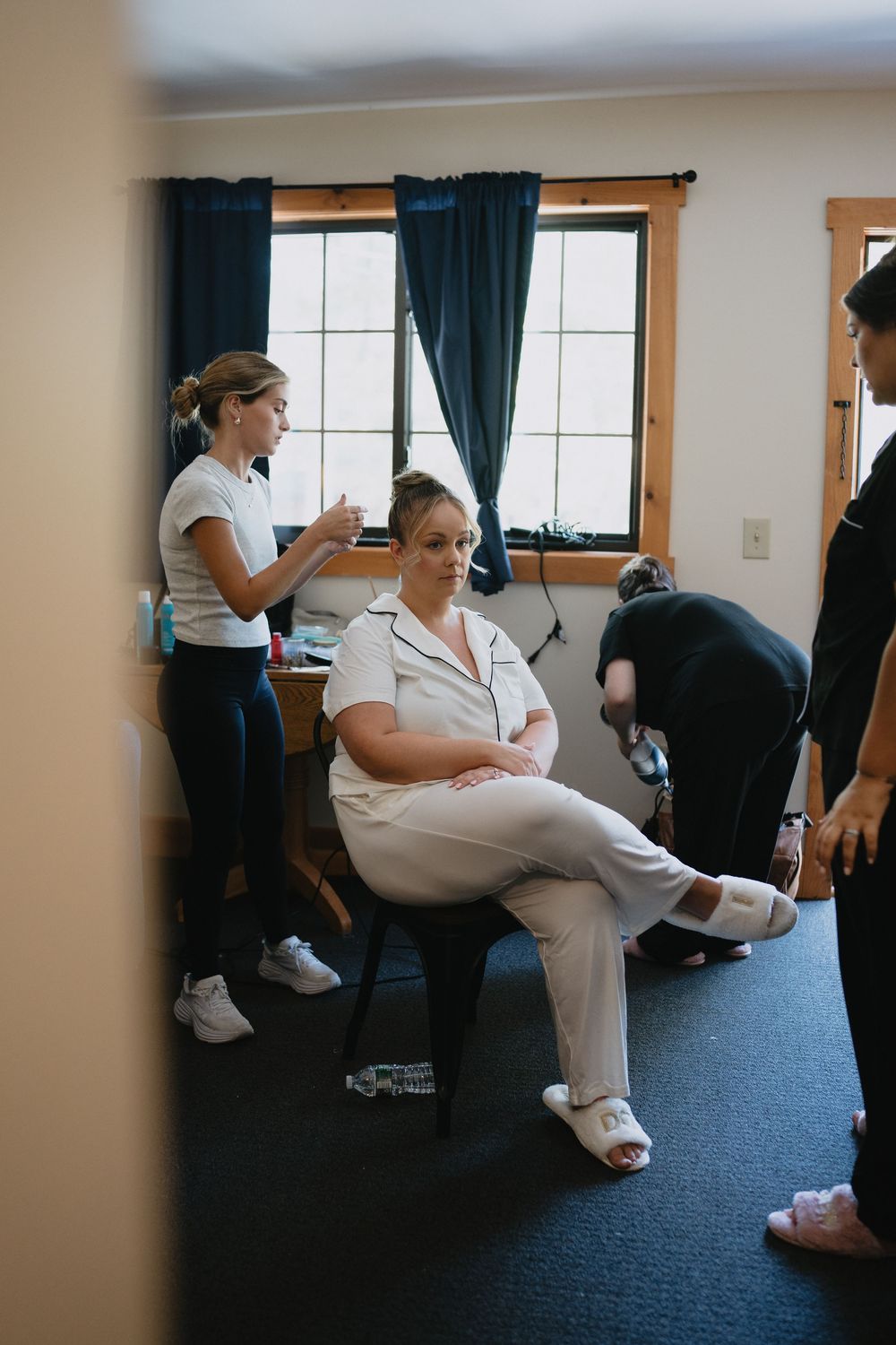 Bride in white robe sits as stylists prepare her hair and accessories in a cozy bridal suite.