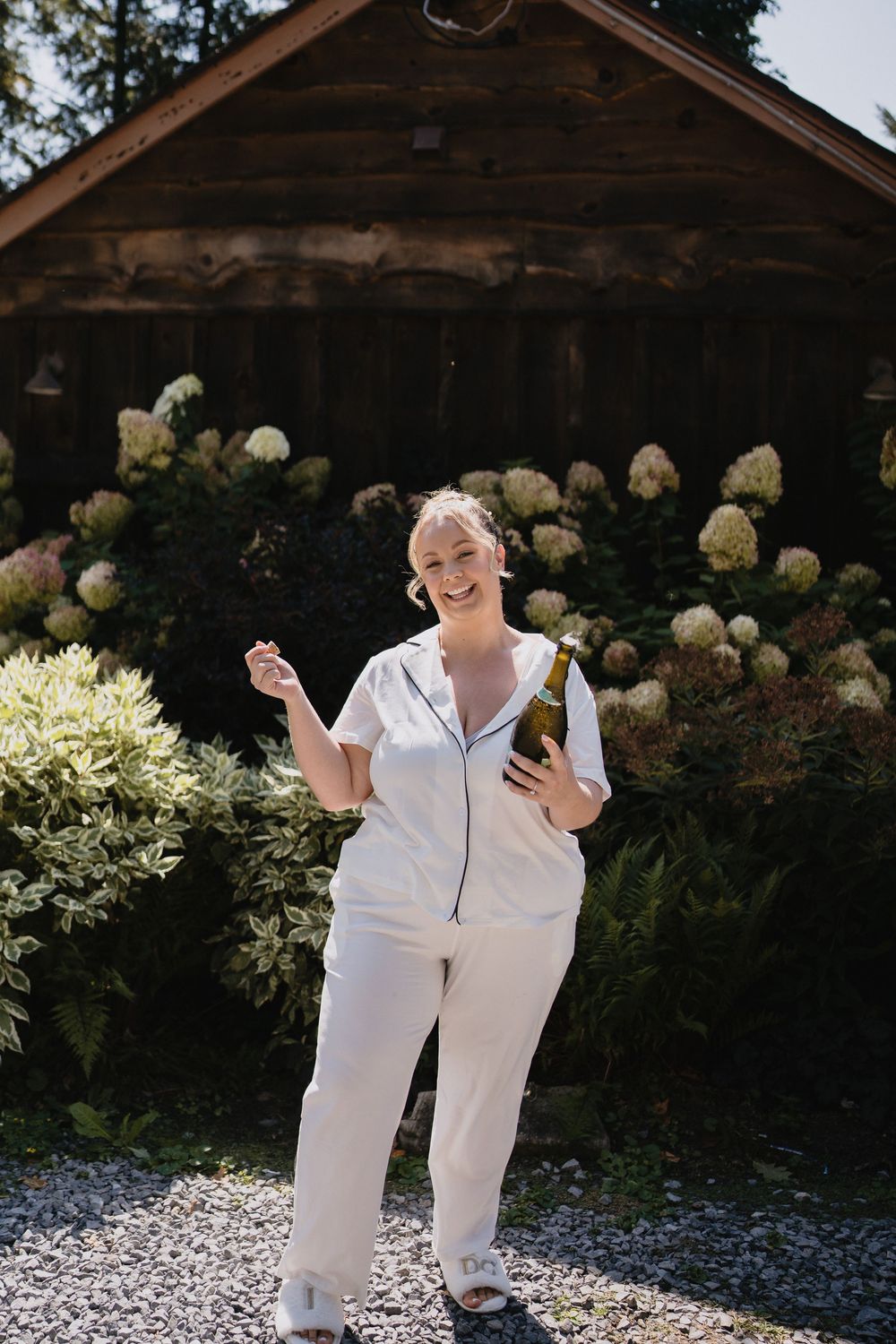 Woman in white outfit holding wine bottle, standing in sunny garden with hydrangeas and wooden cabin backdrop.