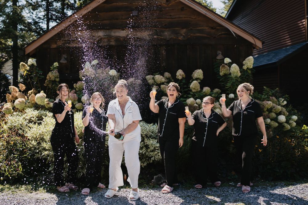 Group of women celebrating outdoors, popping champagne bottle with spray, wearing black dresses and white outfit.