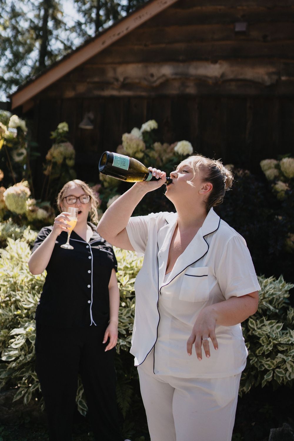 Two women in pajamas celebrate outdoors, one drinking champagne from a bottle near a rustic cabin.