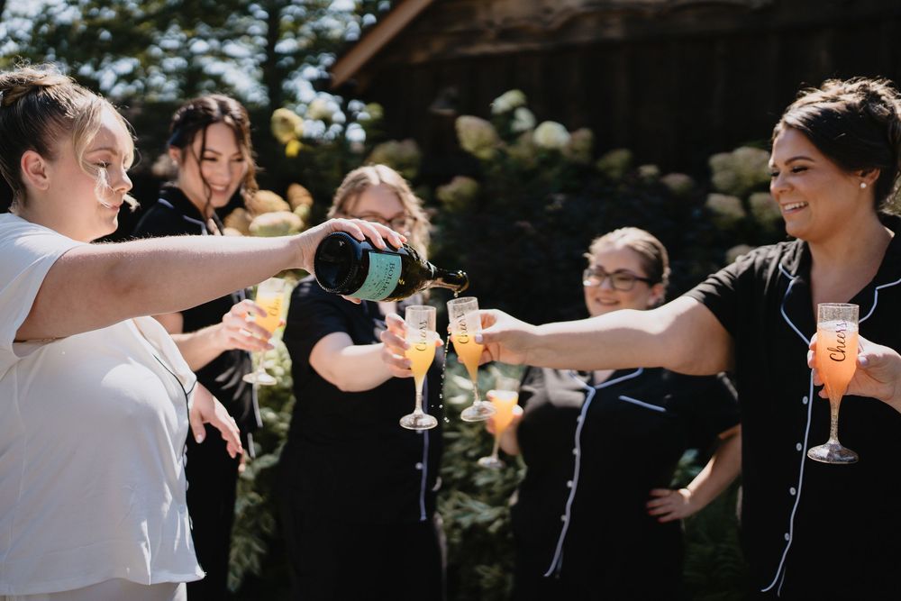 Wedding party toasting with champagne glasses outdoors, bridesmaids in black dresses celebrating with bubbly.
