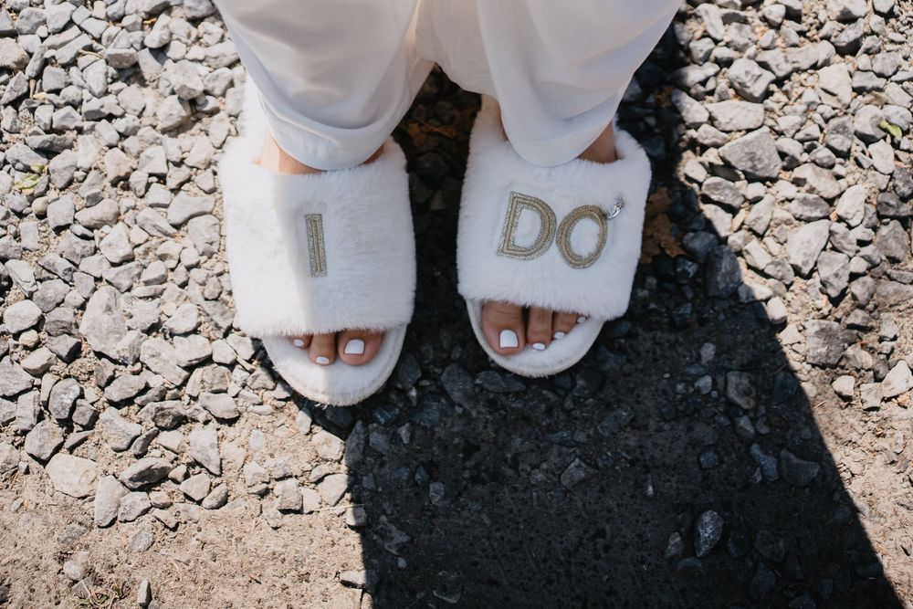 Close-up of bridal feet wearing white fluffy "I DO" slippers on a gravel surface.
