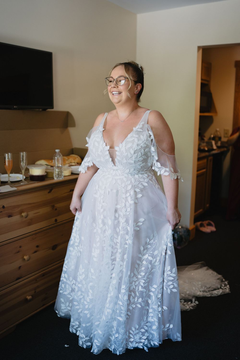 Smiling bride in white floral lace A-line wedding gown with cold shoulders and deep V-neckline, wearing glasses.