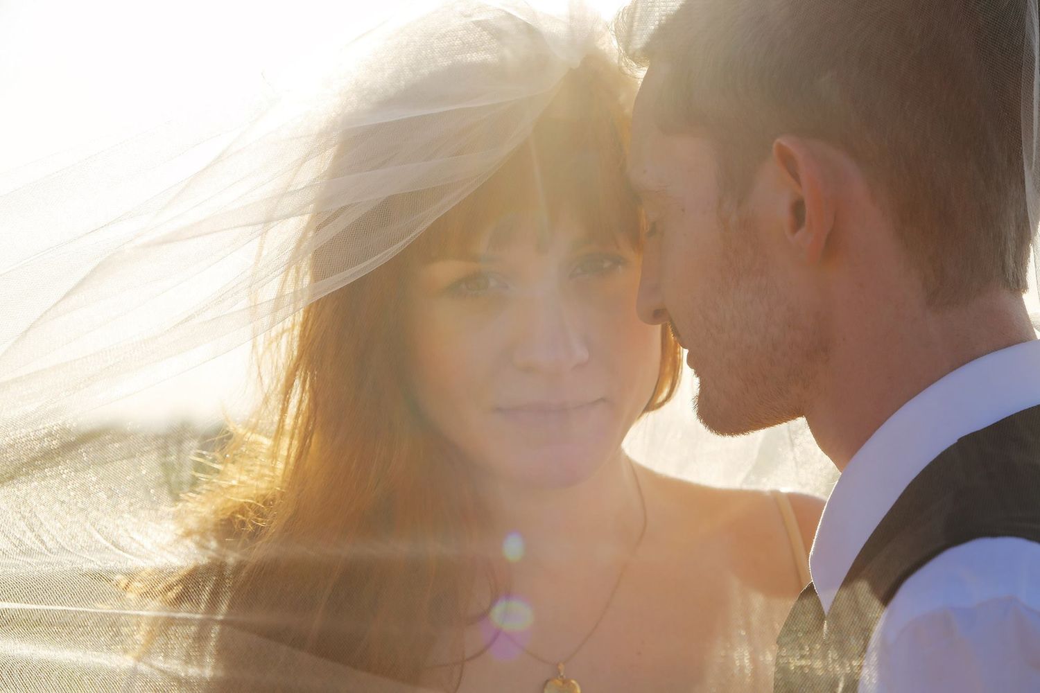 Close-up of a couple touching foreheads in warm golden sunlight with a sheer veil flowing softly around them.
