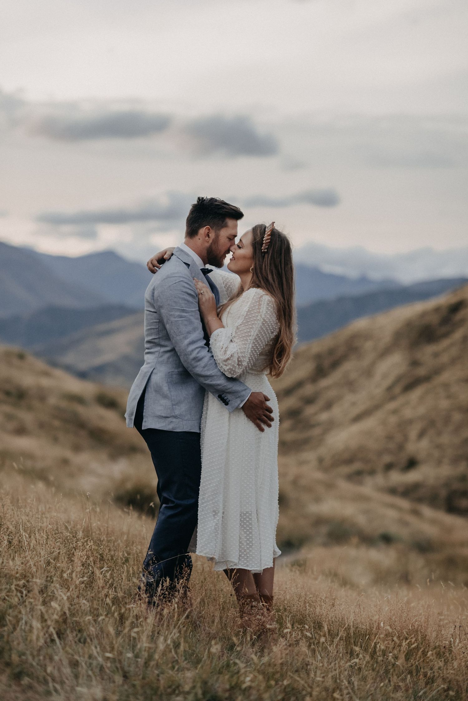 Couple embracing and almost kissing in a golden field with misty mountain backdrop, woman in white dress, man in gray suit.
