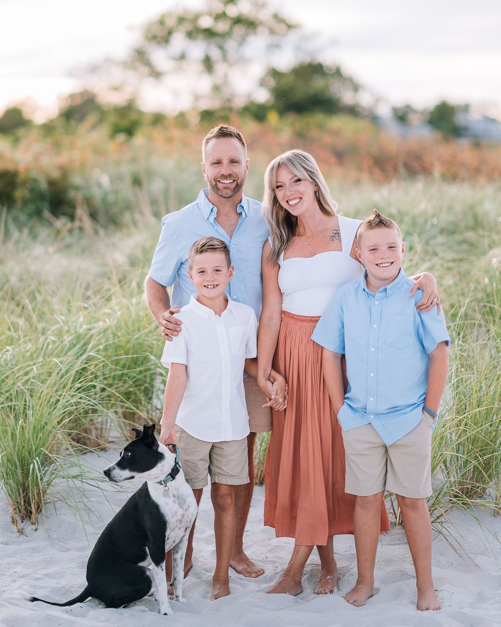 Family of four with a dog posing on a sandy beach with dune grass, wearing casual summer outfits at sunset.