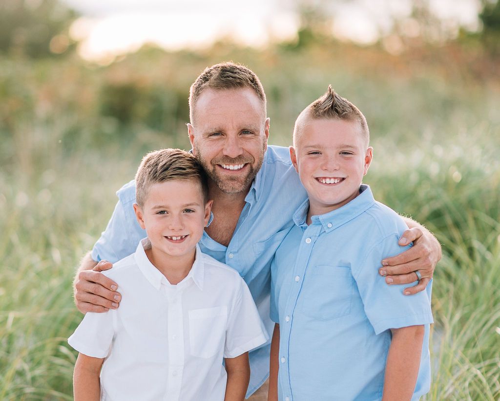 A smiling man with two young boys in blue and white shirts posing outdoors in a grassy field.